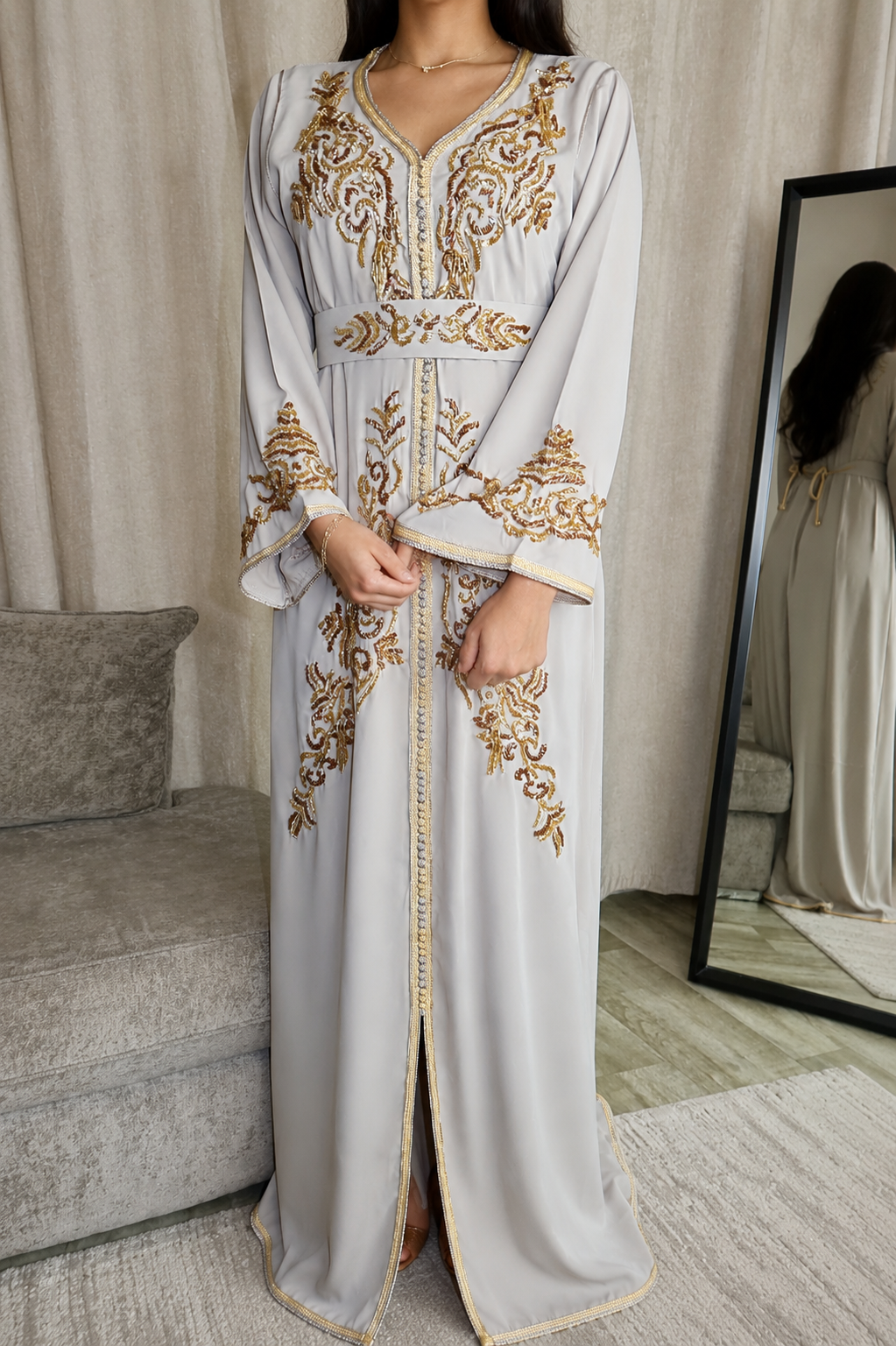 A woman wearing a long light gray abaya with ornate gold embroidery, standing in a room with a gray sofa and a mirror showing her reflection.