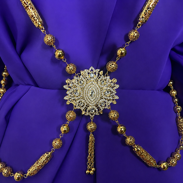 Traditional Moroccan Hamela gold chain featuring intricate filigree detailing, crystal centerpiece accents, and elegant draped chains styled on rich purple fabric for luxury Moroccan caftan and occasion wear.
