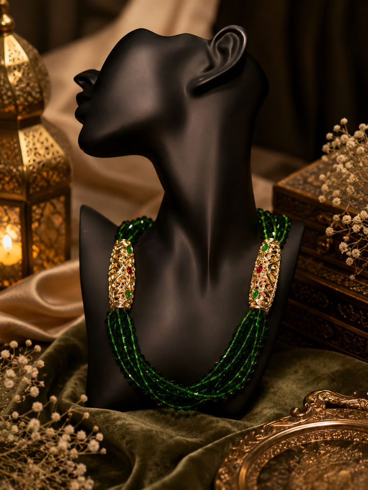 Luxury Moroccan-inspired green beaded necklace displayed on a matte black mannequin bust, featuring layered emerald crystal strands with ornate gold filigree accents and red-green gemstone detailing in an elegant warm-toned setting.
