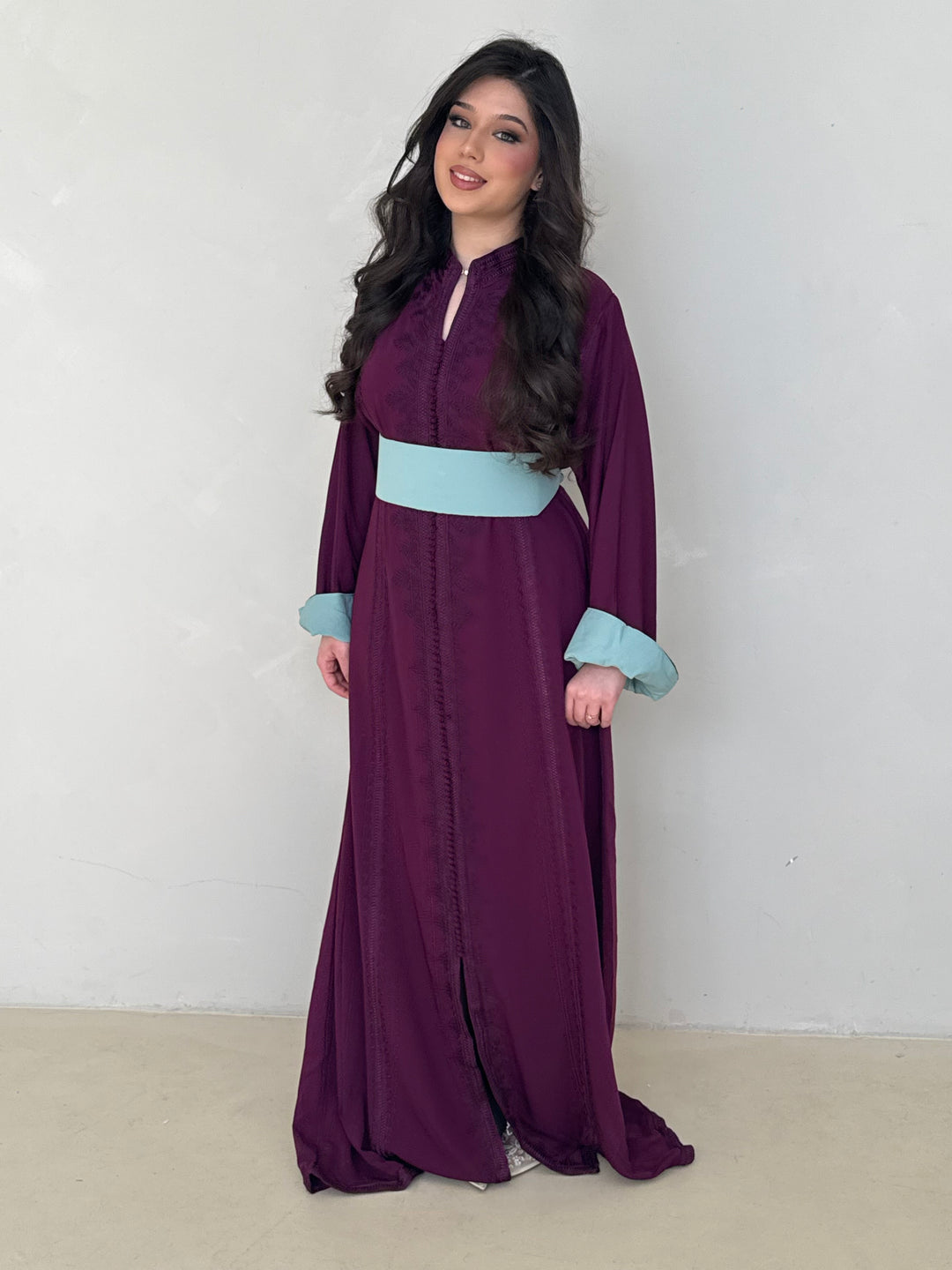 Woman wearing a long purple modest caftan dress with a light blue belt against a white background
