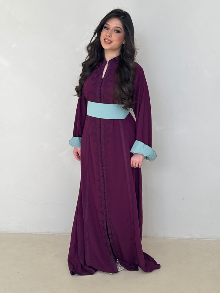 Woman wearing a long purple modest caftan dress with a light blue belt against a white background