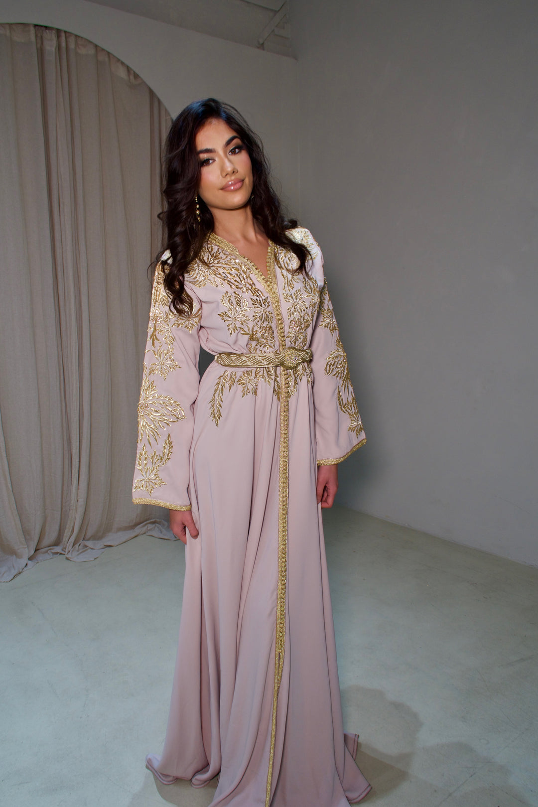 Woman wearing a long, light-colored dress with gold embroidery in a minimalistic setting.